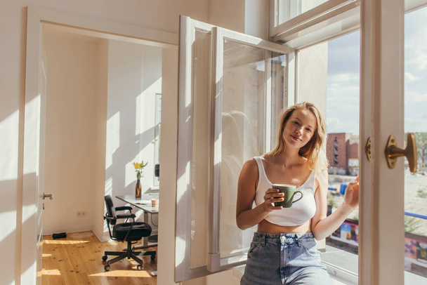 Smiling woman in top holding cup near open window at home  - Fotoğraf, Görsel