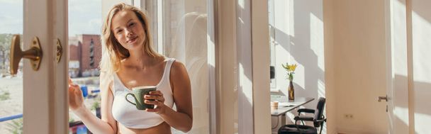 Blonde woman in top holding cup of coffee near open window at home, banner  - Photo, Image