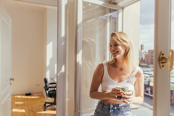 Cheerful young woman in top holding cup near open window at home  - Foto, Bild