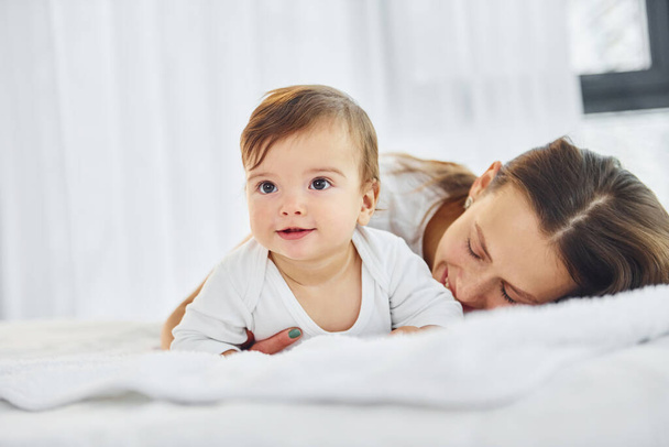 On the white bed. Mother with her little daughter is indoors at home together. - Photo, Image