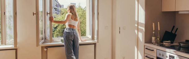 Young woman looking at open window near cup on windowsill in kitchen, banner  - Фото, зображення