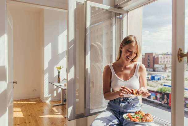 Smiling woman holding croissant while sitting on windowsill at home  - Photo, image