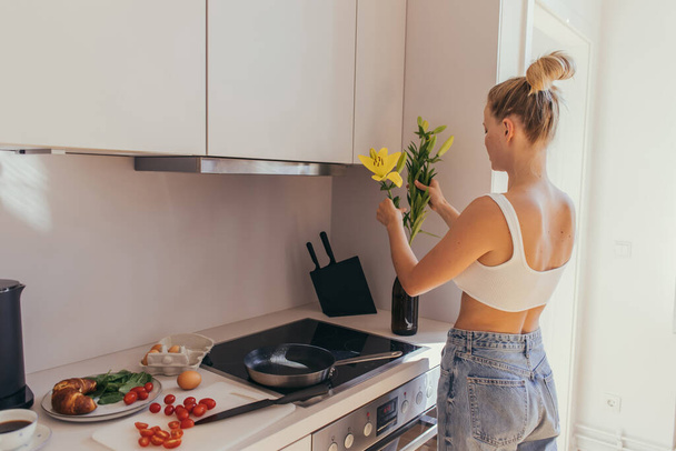 Blonde woman putting flowers in vase near breakfast and coffee in kitchen  - Фото, изображение