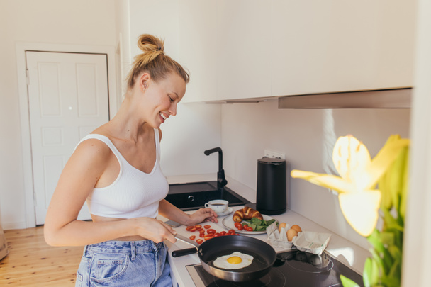 Side view of smiling blonde woman looking at egg on frying pan near coffee and cherry tomatoes in kitchen  - Photo, Image