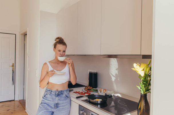 Mujer joven en la parte superior sosteniendo taza de café cerca del desayuno y flores de lirio en la cocina  - Foto, imagen