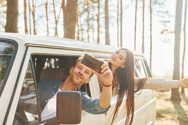 Making selfie. Young couple is traveling in the forest at daytime together. - Фото, изображение