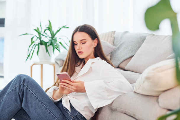 Sits near sofa with phone in hands. Young woman in white shirt and jeans is at home. - Foto, immagini