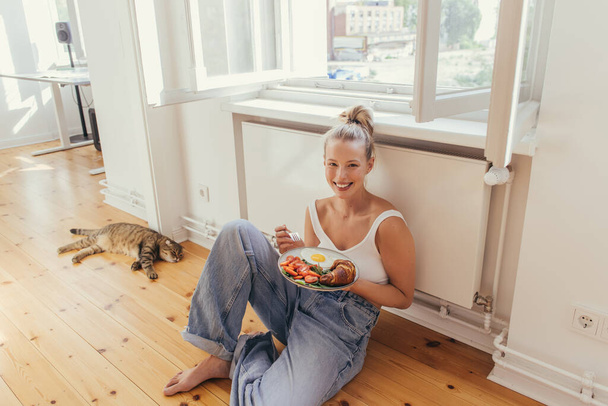 Positive young woman holding plate with delicious breakfast near scottish fold cat at home  - Fotó, kép