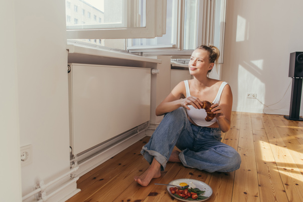 Smiling woman holding croissant near open window and breakfast at home  - Foto, Imagem