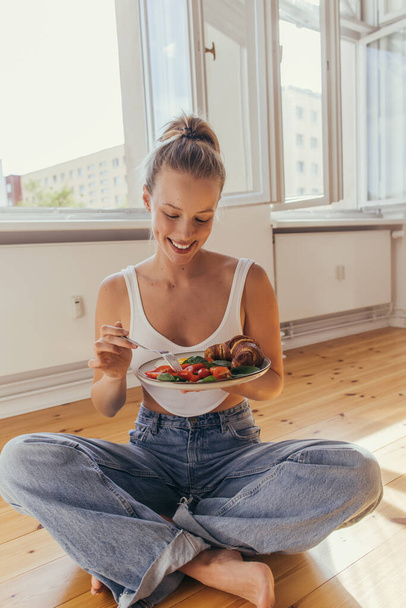 Positive woman in top and jeans holding plate with tasty breakfast on floor at home  - Foto, immagini