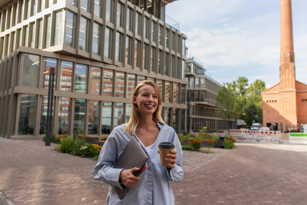 Smiling freelancer holding coffee to go and laptop on urban street in Berlin  - Φωτογραφία, εικόνα