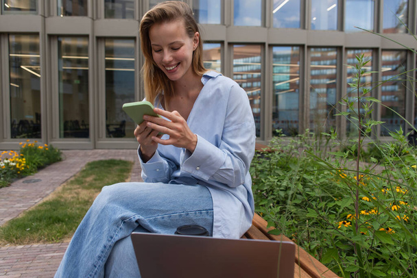 Smiling freelancer using smartphone near laptop on bench outdoors  - Fotó, kép