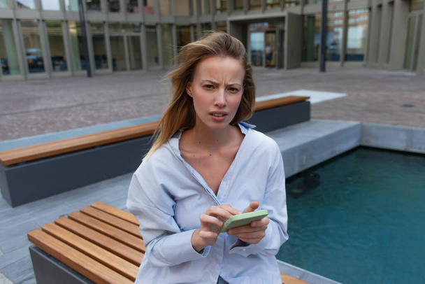 Huolestunut nainen paita tilalla älypuhelin ja katsot kameraa kaupunkien kadulla  - Valokuva, kuva