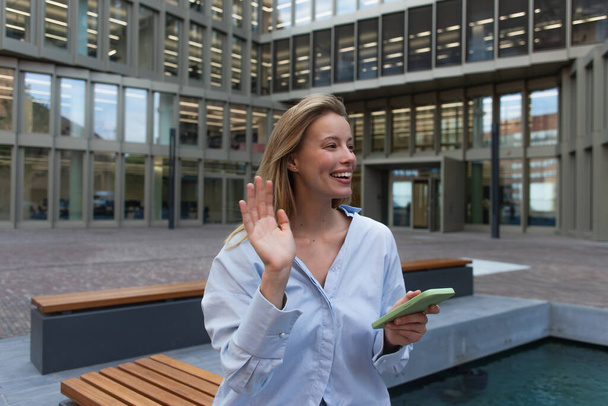 Happy blonde woman in shirt holding smartphone and waving hand on urban street  - Fotografie, Obrázek