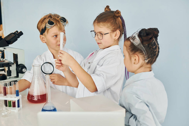 Standing by the table with test tubes. Children in white coats plays a scientists in lab by using equipment. - Φωτογραφία, εικόνα
