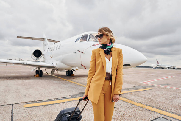 In yellow clothes. Young woman with luggage is outdoors near airplane. - 写真・画像