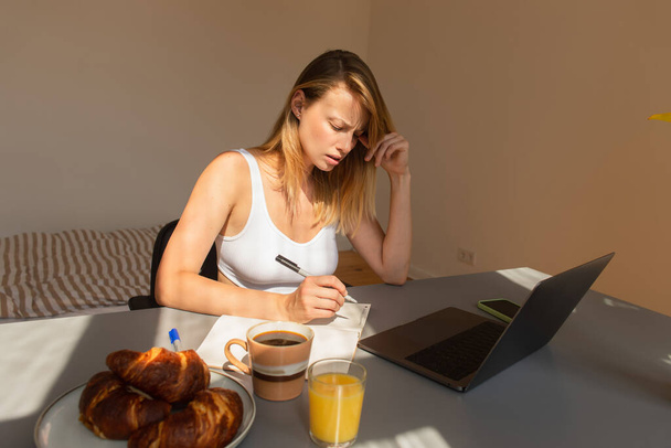 Worried freelancer writing on notebook near devices and breakfast on table at home  - Foto, Imagen