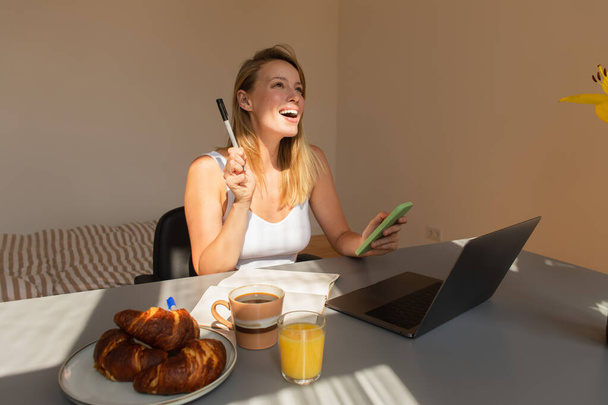 Excited freelancer holding smartphone and pen near laptop and breakfast at home  - Valokuva, kuva