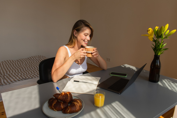 Smiling freelancer holding cup near breakfast and devices at home  - Φωτογραφία, εικόνα