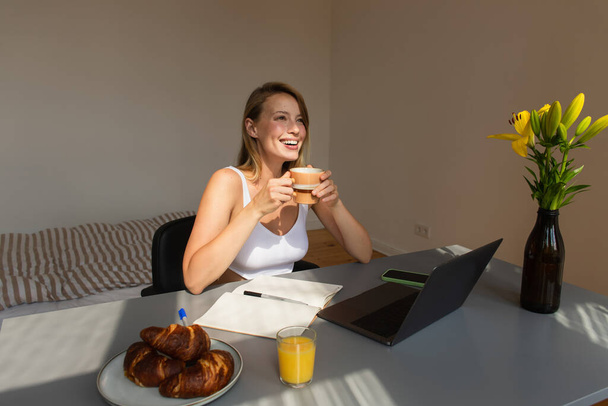 Happy blonde woman holding cup near breakfast and gadgets at home  - Foto, Imagem