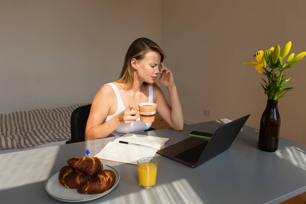 Side view of freelancer holding cup while suffering from headache near devices and breakfast at home  - Fotografie, Obrázek
