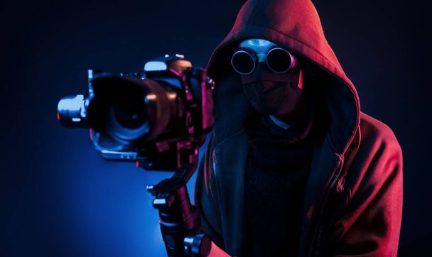Guy in hood with camera. Neon lighting. Young european man is in the dark studio. - Foto, Imagen