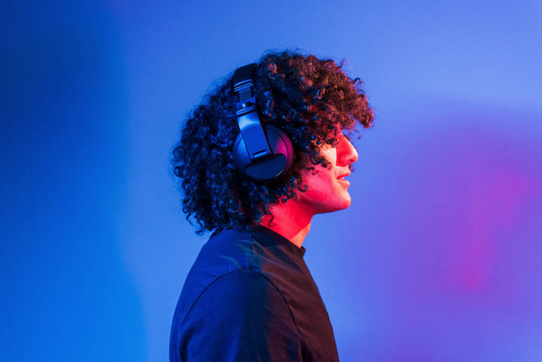 With headphones. Young beautiful man with curly hair is indoors in the studio with neon lighting. - Fotó, kép