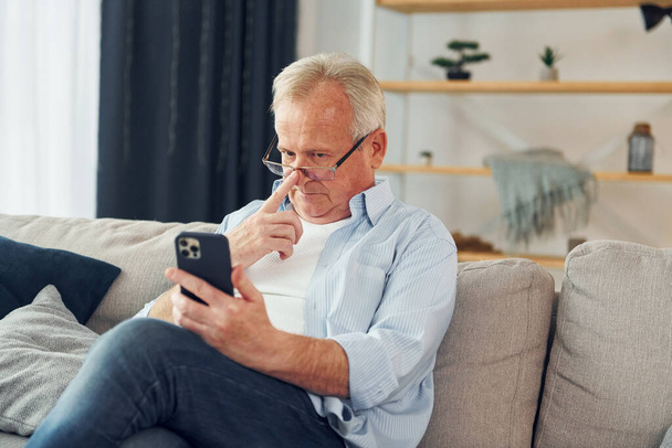 Sitting with smartphone in hands. Senior man in nice clothes is at home. - Fotoğraf, Görsel