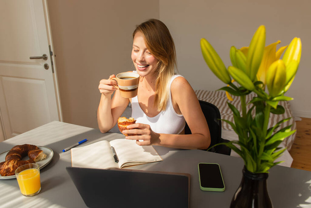 Fröhliche Freiberuflerin mit Kaffee und Croissant in der Nähe von Gadgets und Blumen zu Hause  - Foto, Bild