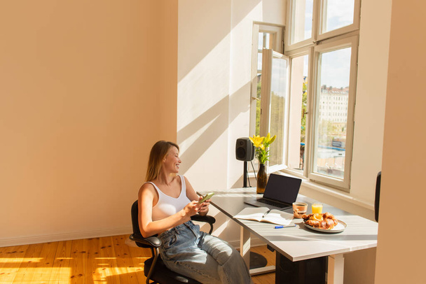 Side view of smiling freelancer using smartphone near laptop and breakfast at home  - Foto, Imagen