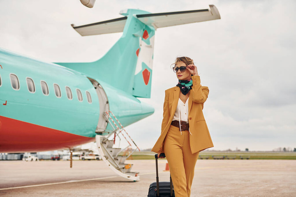 Passenger woman that is in yellow clothes, sunglasses and with luggage is outdoors near plane. - Foto, Imagem