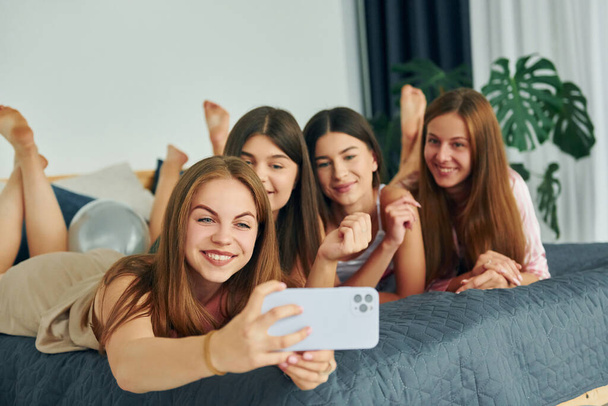 Laying down on bed with smartphone. Group of happy women that is at a bachelorette party. - Photo, image