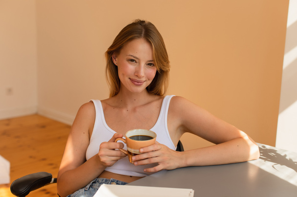 Sarışın kadın elinde kahve fincanıyla evdeki defterin yanındaki kameraya bakıyor.  - Fotoğraf, Görsel