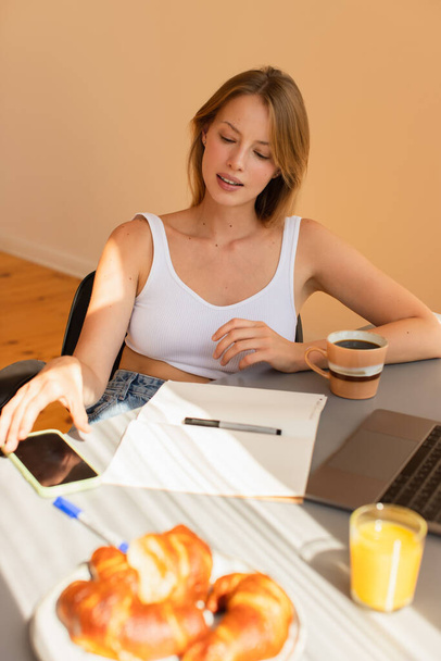 Blonde freelancer in top taking smartphone near notebook and blurred breakfast at home  - Photo, Image