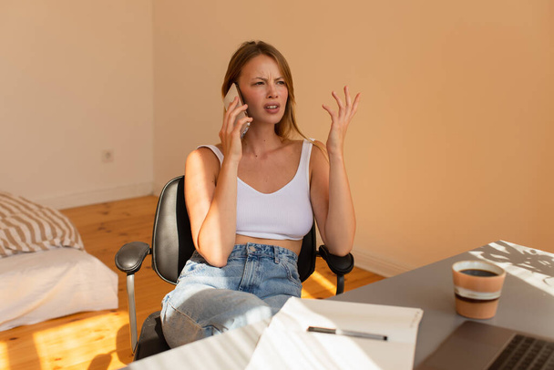 Stresszes szabadúszó beszél mobilon közel laptop és kávé otthon  - Fotó, kép