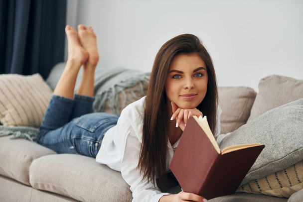 With book. Beautiful young woman in jeans and white shirt is at home. - Fotoğraf, Görsel