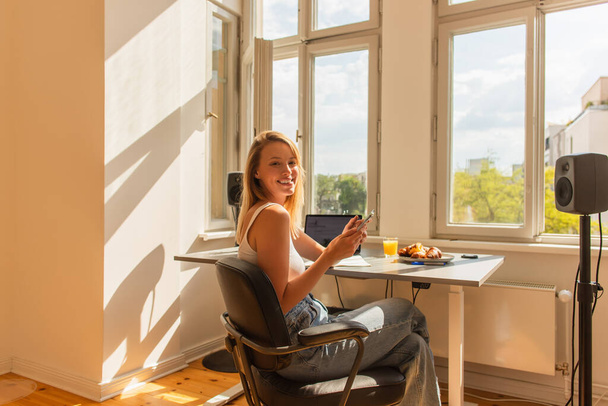 Feliz rubia freelancer utilizando el teléfono celular cerca de zumo de naranja y desayuno en casa  - Foto, Imagen