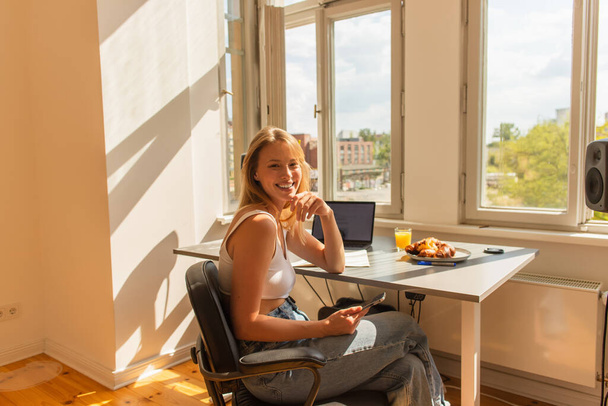 Positive blonde freelancer looking at camera near devices and breakfast at home  - Fotografie, Obrázek