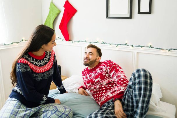Pareja feliz vistiendo pijamas y suéteres de Navidad relajándose en la cama juntos en la mañana  - Foto, Imagen
