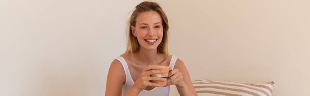 Jonge blonde vrouw met kopje koffie en kijken naar de camera thuis, banner  - Foto, afbeelding