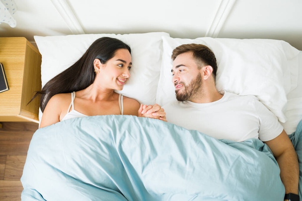 Top view of a beautiful couple in love holding hands and cuddling under the covers in bed while waking up together  - Photo, Image