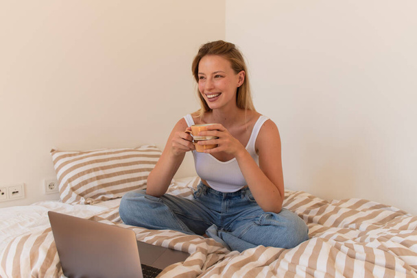 Щаслива блондинка тримає чашку кави біля ноутбука на ліжку
  - Фото, зображення