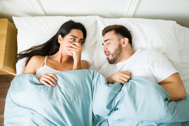 Femme latine bouleversée couvrant son nez à cause de la mauvaise haleine de son partenaire endormi au lit - Photo, image