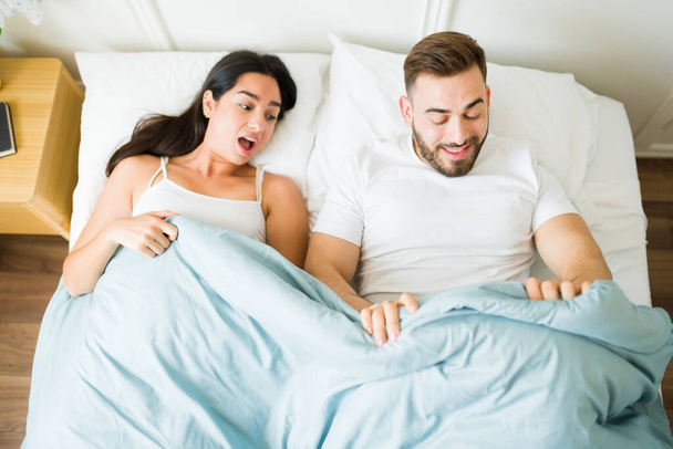 Meglepett fiatal nő néz a takaró alá egy megdöbbent férfi partnerrel, és készen áll a szexre a hálószobában - Fotó, kép