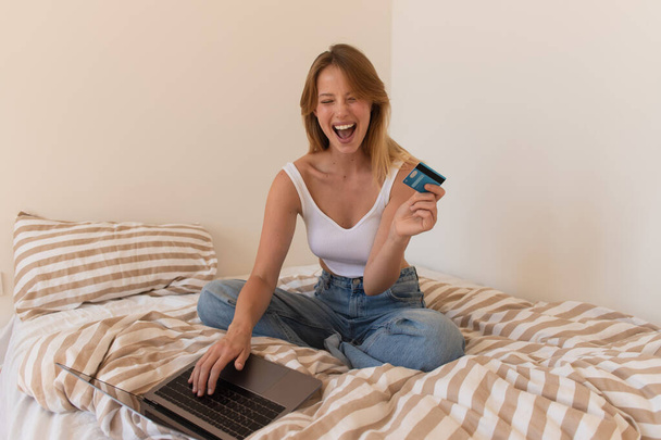 Kredi kartı tutan ve yatağında dizüstü bilgisayar kullanan heyecanlı genç bir kadın.  - Fotoğraf, Görsel