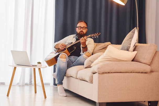 Feeling relaxed. Man in casual clothes and with acoustic guitar is indoors. - 写真・画像