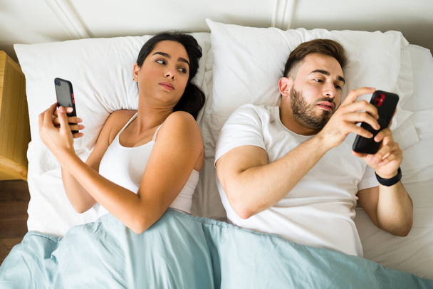 Jealous hispanic woman in bed spying on his happy partner or husband while texting on the phone and cheating - Photo, Image