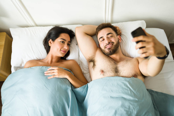 Όμορφος νεαρός άνδρας που βγάζει μια σέλφι με το σύντροφό του ή τη φίλη του κάτω από τα σκεπάσματα για να την ανεβάσει στα μέσα κοινωνικής δικτύωσης αφού κοιμηθεί μαζί - Φωτογραφία, εικόνα