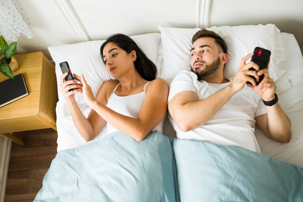 Расстроенный кавказский мужчина шпионит за своей девушкой или женой, когда пишет смс на смартфоне, изменяя и отдыхая в постели - Фото, изображение
