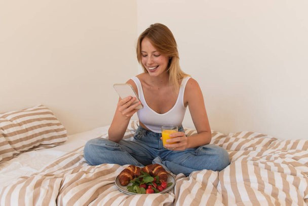 Улыбающаяся женщина с помощью смартфона и держа апельсиновый сок возле завтрака на кровати  - Фото, изображение